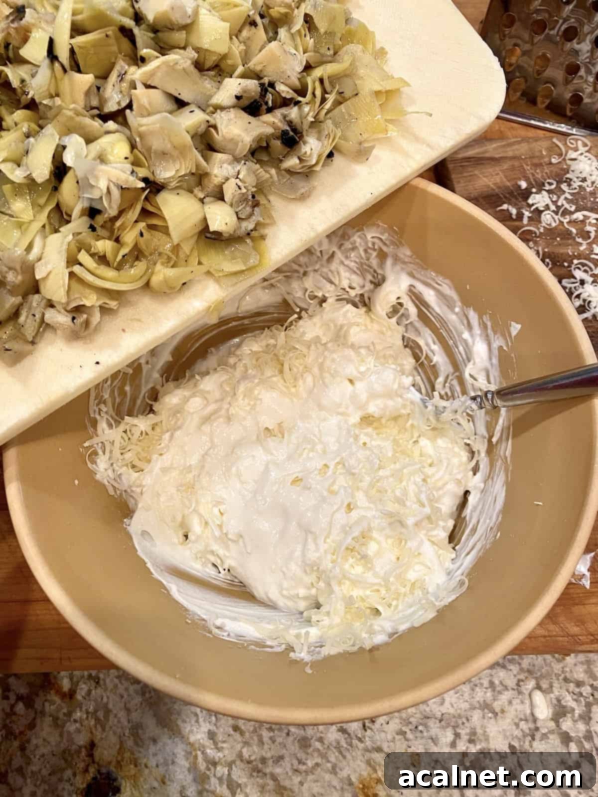A bowl of creamy, unmixed ingredients for artichoke dip, with a cutting board of freshly chopped artichoke hearts poised nearby.