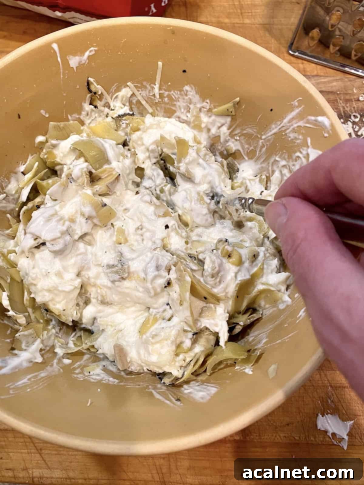 Creamy dip base with cheese and chopped artichoke hearts added, ready for a gentle stir.