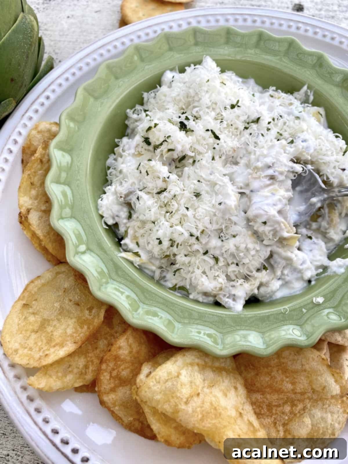 A small bowl of creamy artichoke dip presented on a cozy side table, accompanied by a handful of chips and crackers.