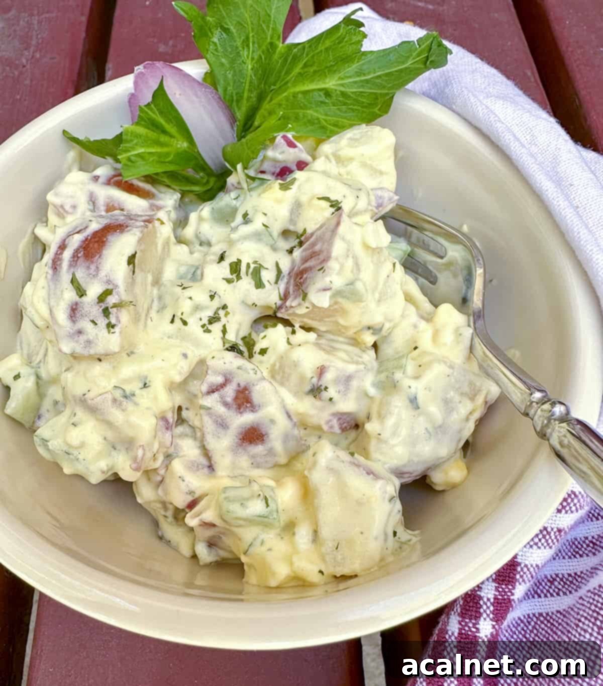 A bowl of creamy red potato salad, garnished with a sprig of celery, a fork resting in the bowl, and a napkin on the side, inviting you to dig in.
