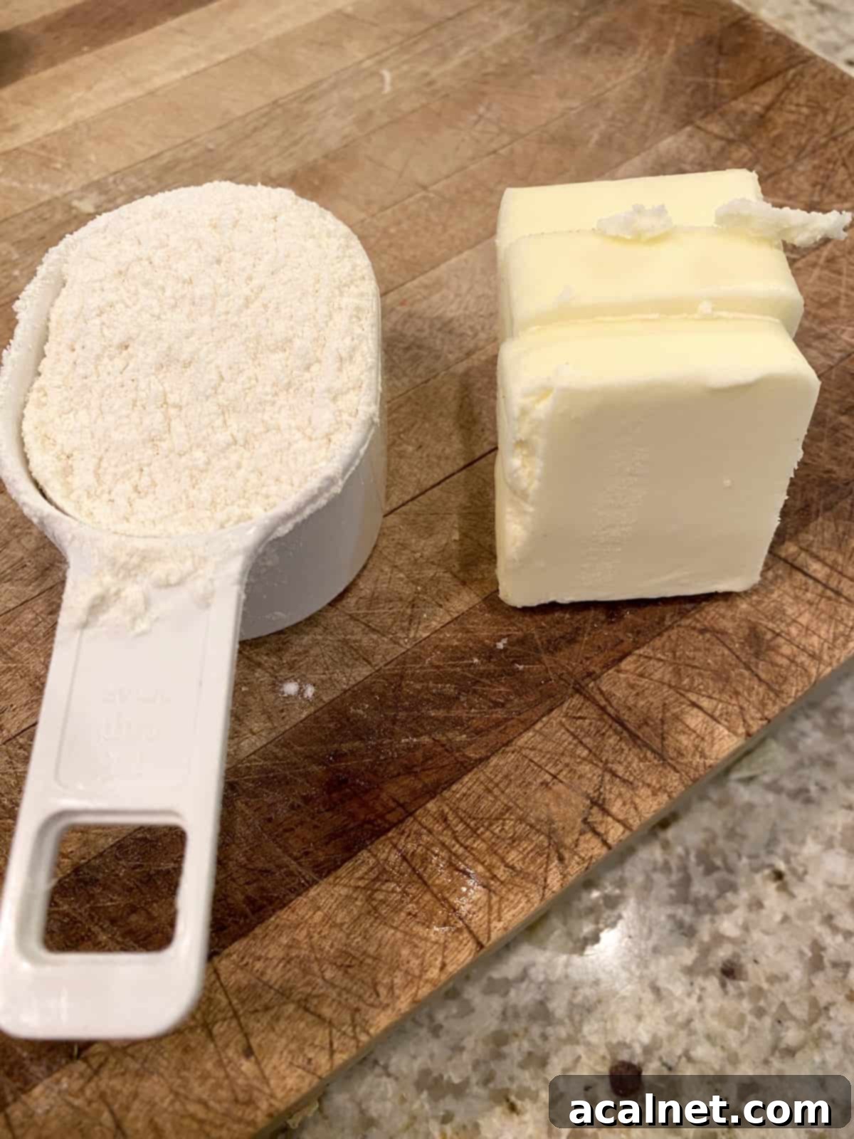 A measuring scoop filled with white flour and a stick of butter on a cutting board, prepared for making a roux.