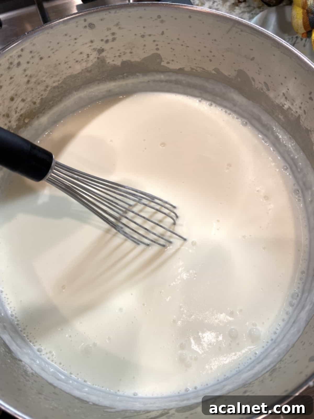 A creamy white sauce simmering in a saucepan on the stove, with a metal whisk resting on the side, indicating it's freshly made.