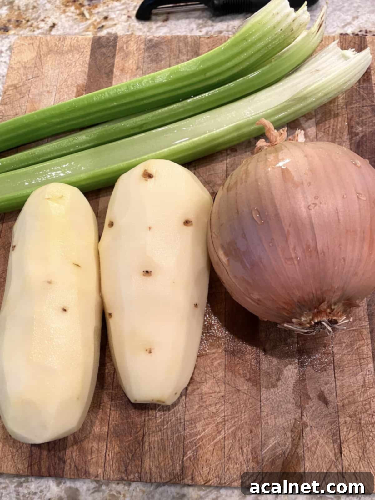 Peeled Russet potatoes, fresh celery stalks, and a brown onion arranged on a wooden cutting board, ready for chopping.