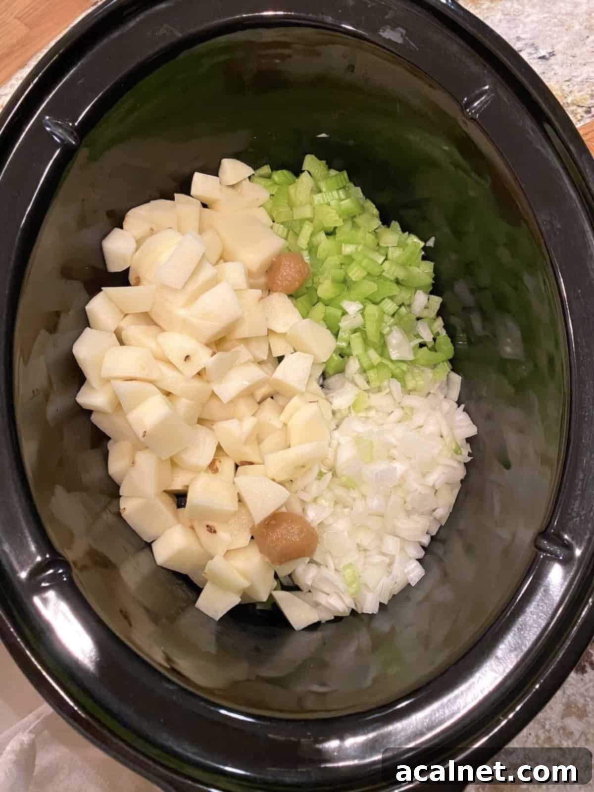 A view inside a slow cooker, showing raw diced vegetables like potatoes, celery, and onions, without the clam juice yet added.