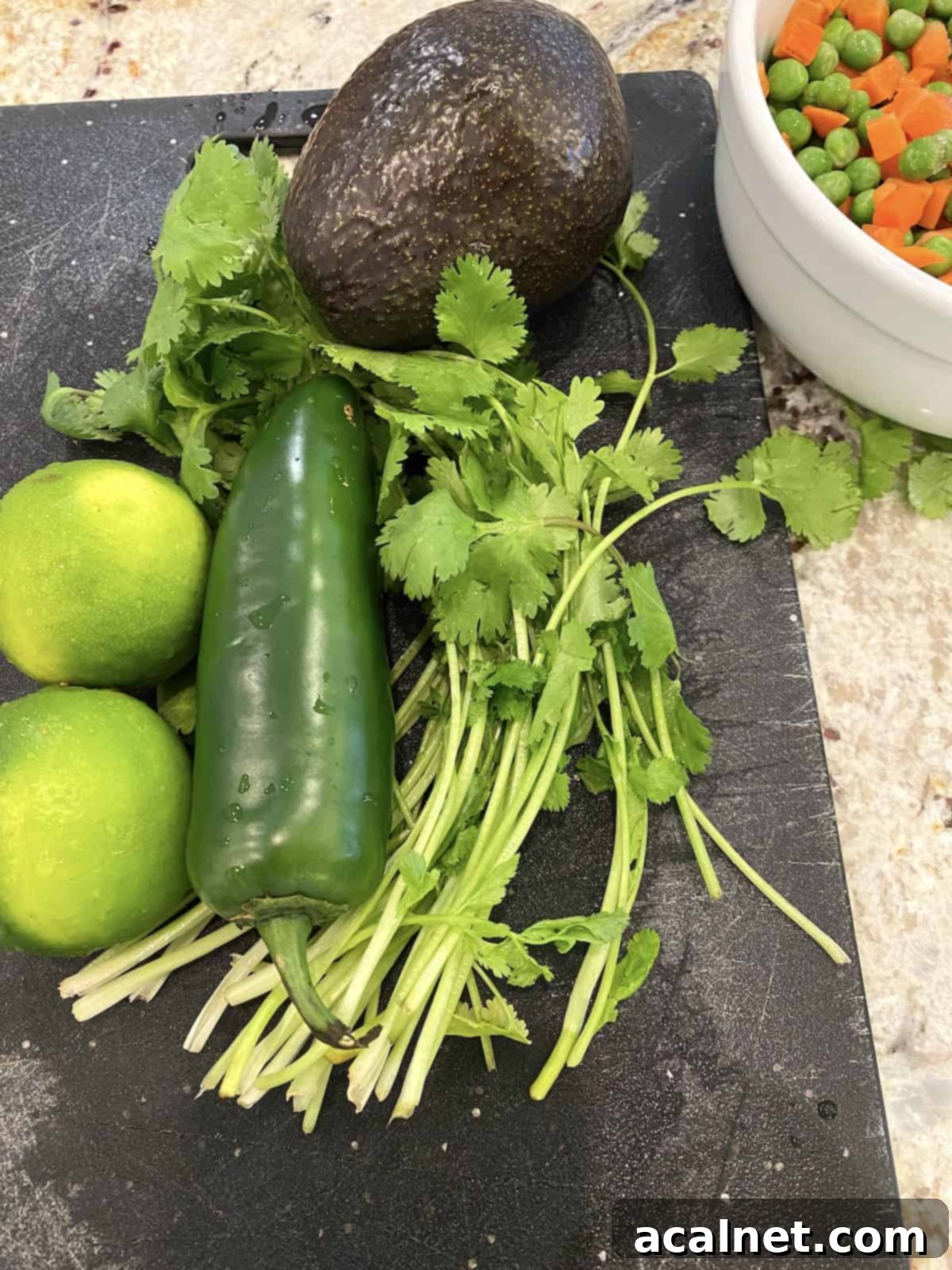 A jalapeno pepper with fresh limes and cilantro on a cutting board next to a bowl of peas and carrots.