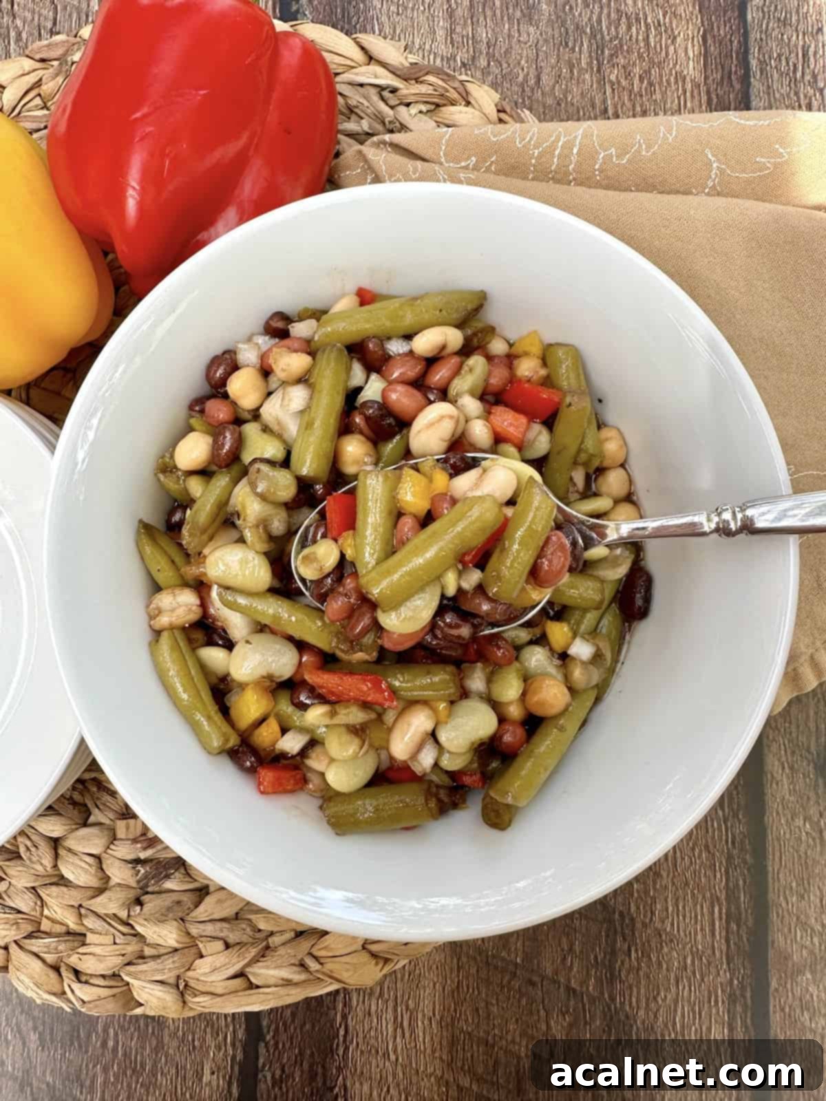 A serving bowl of cold green bean salad with a spoon, on a place mat with two peppers off to the side.