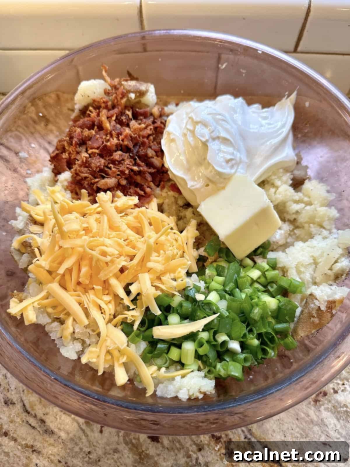Cheese, green onions, cooked bacon, sour cream and butter arranged on top of mashed, baked potatoes in a bowl.