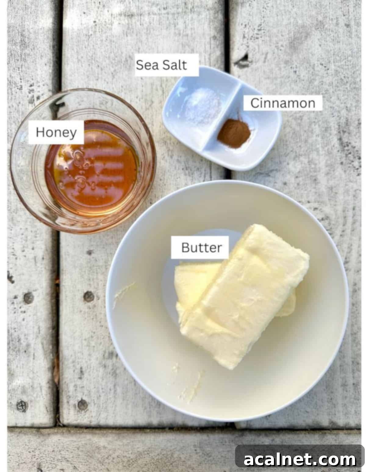 Ingredients for honey butter, including sticks of softened butter, pure honey, ground cinnamon, and fine sea salt, arranged in small bowls on a rustic wooden table.