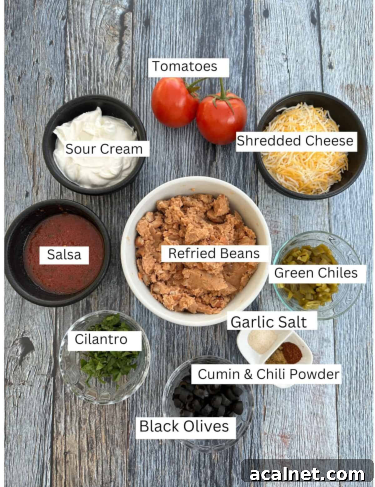 Labeled ingredients for five-layer taco dip on a wooden table, including beans, sour cream, cheese, salsa, and toppings.