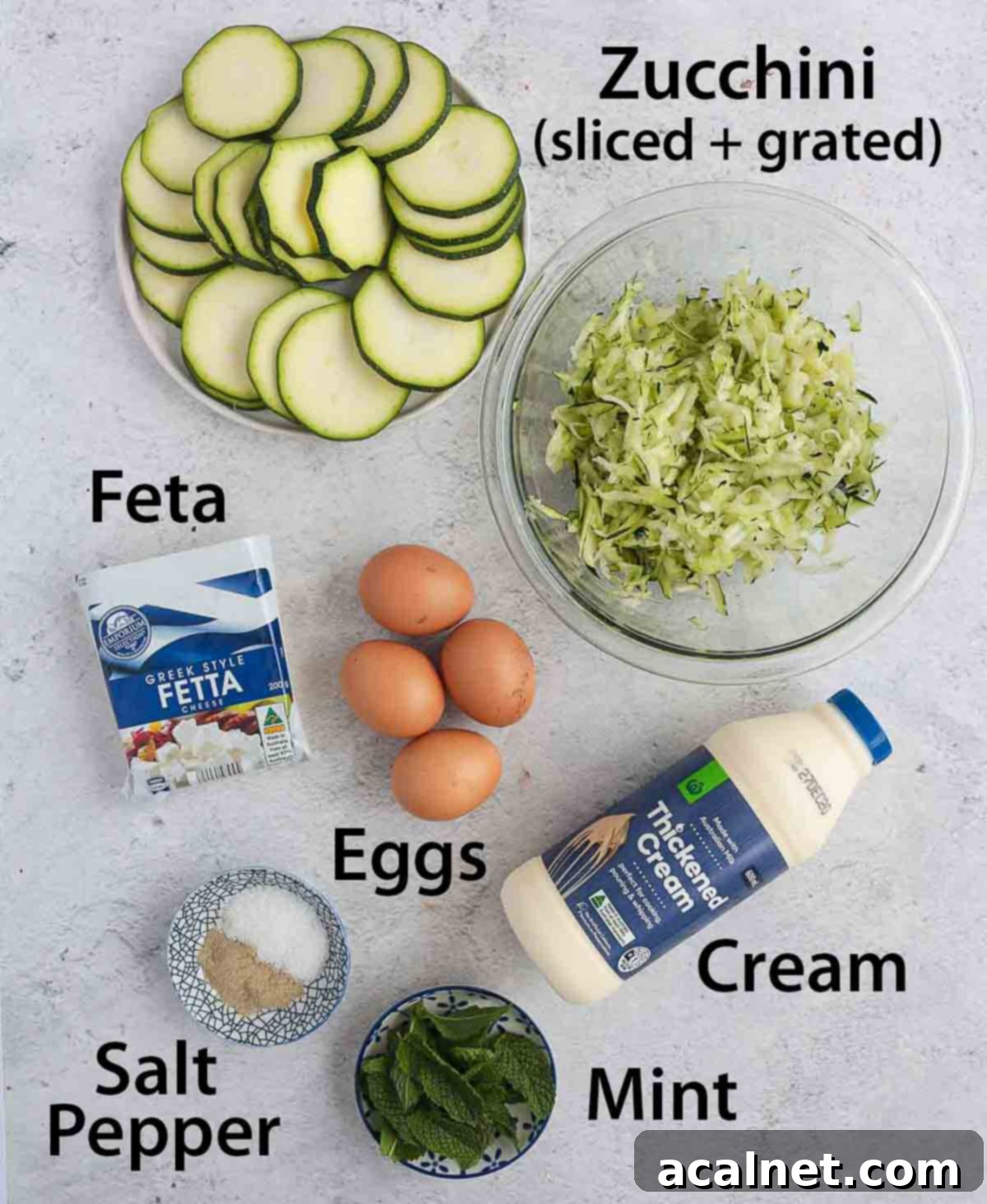 A collection of fresh ingredients for Zucchini Quiche, including zucchini, eggs, cream, feta, and mint, neatly arranged on a grey surface.