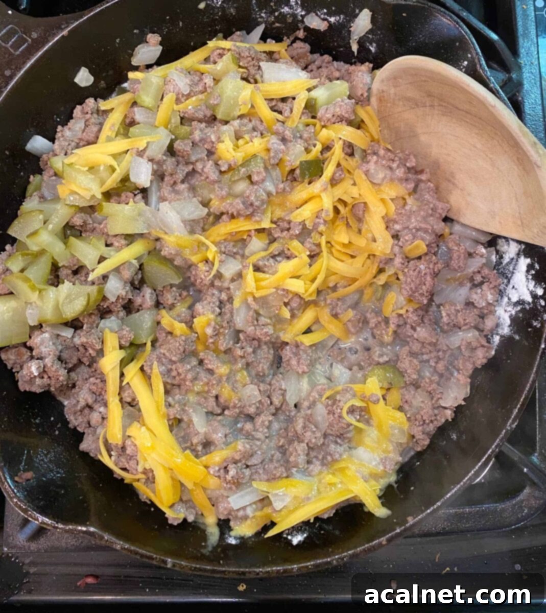 Fried ground beef mixed with shredded cheese and diced pickles in a cast iron skillet on a stove, forming the rich cheeseburger pie filling.