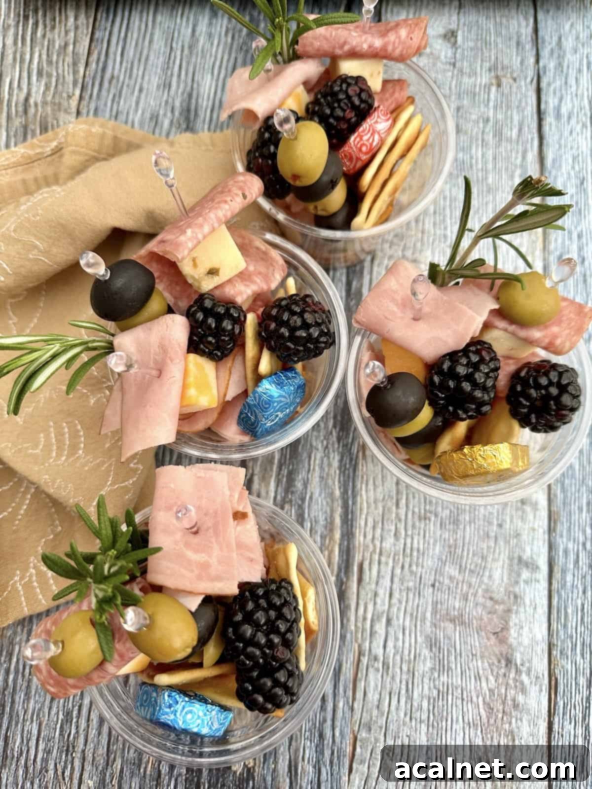 Four charcuterie cups on a plank with a cloth napkin.