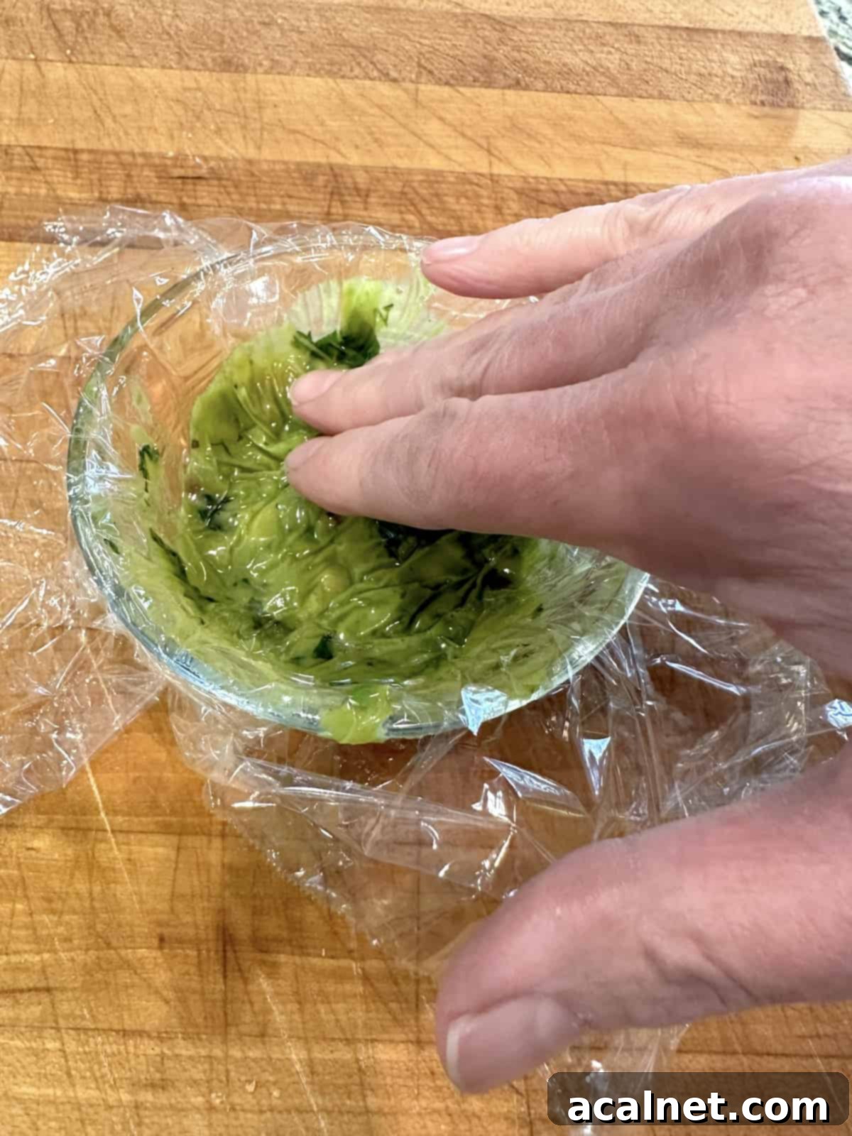 A hand pushing plastic wrap on the surface of guacamole in a bowl.