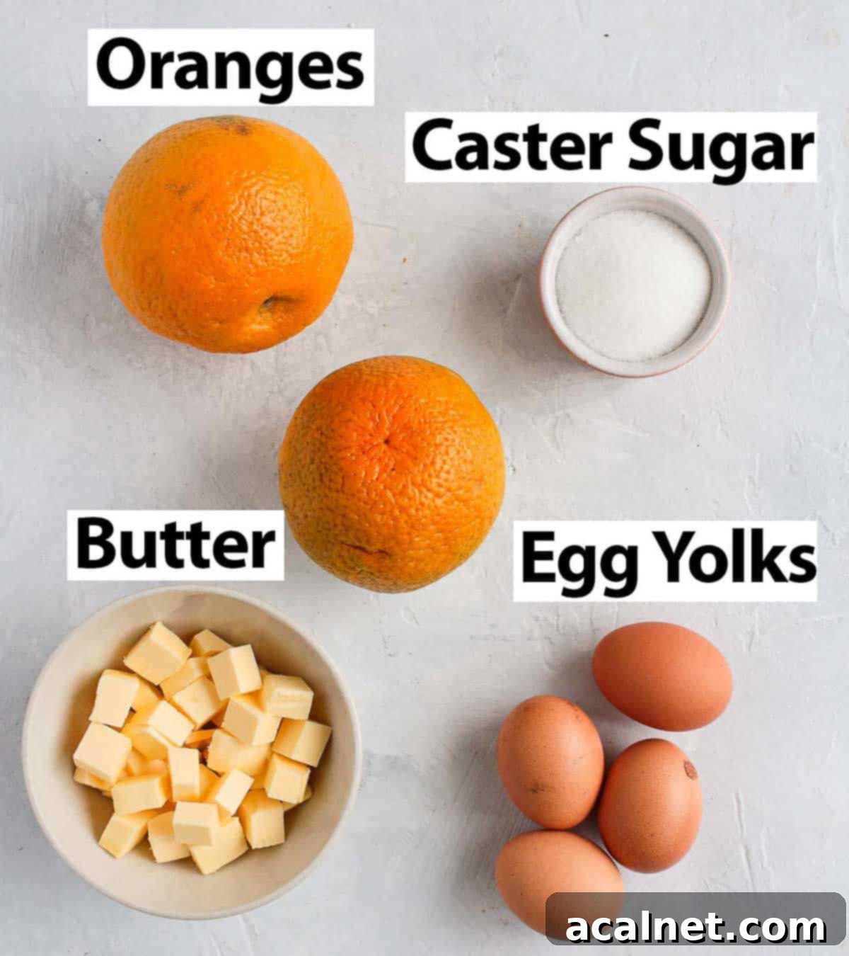 Fresh ingredients laid out on a white surface, including vibrant oranges, a bowl of caster sugar, cubed butter, and separated egg yolks.
