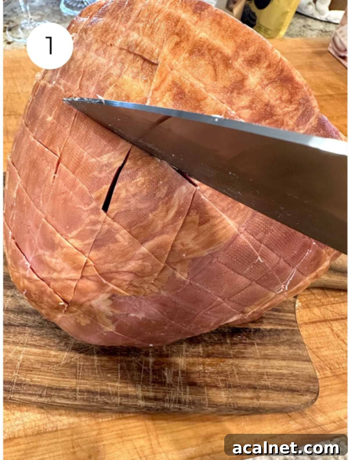 A fully cooked ham on a cutting board, being scored with a sharp knife in a criss-cross pattern.