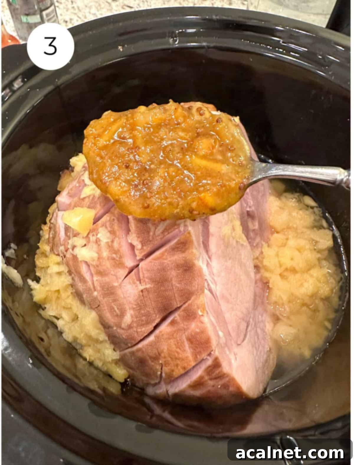 A spoon applying the marmalade-mustard glaze over a ham placed in a slow cooker, with crushed pineapple already at the bottom.