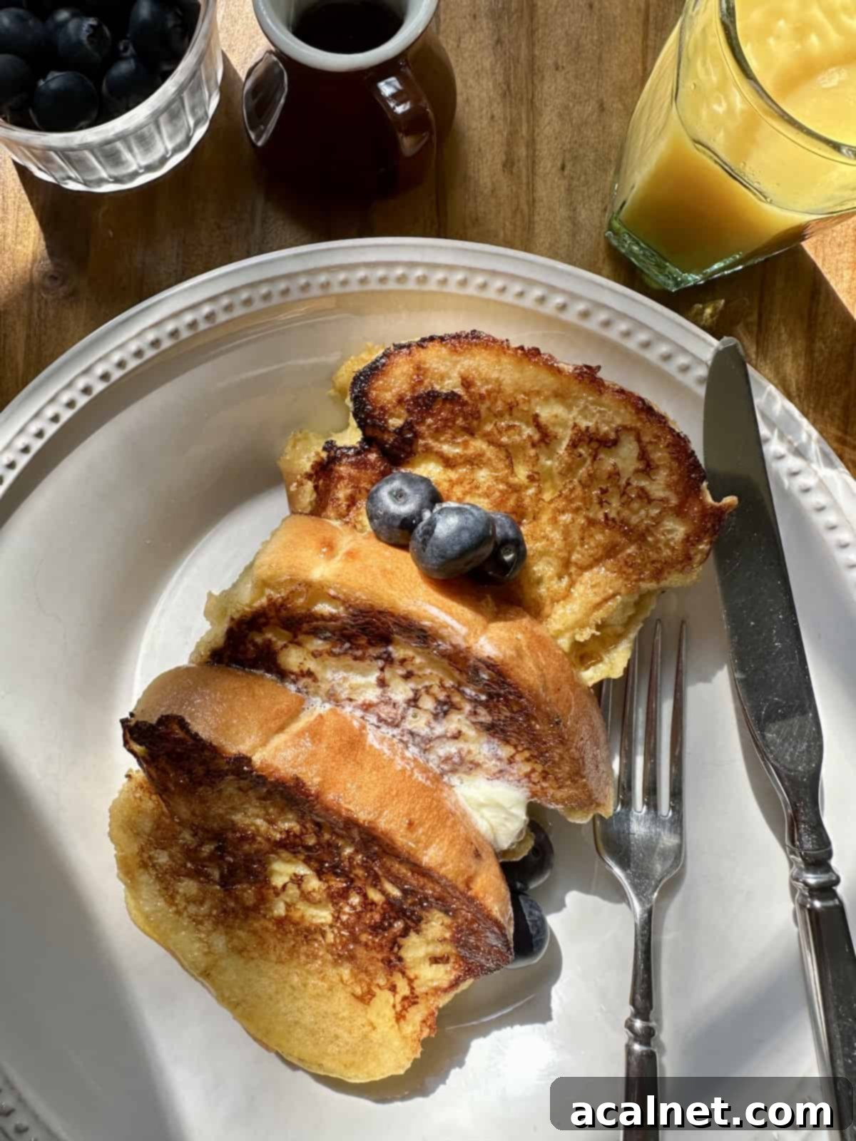 Several slices of buttermilk french toast on a plate with an abundance of fresh berries, a glass of orange juice, and a pitcher of maple syrup.
