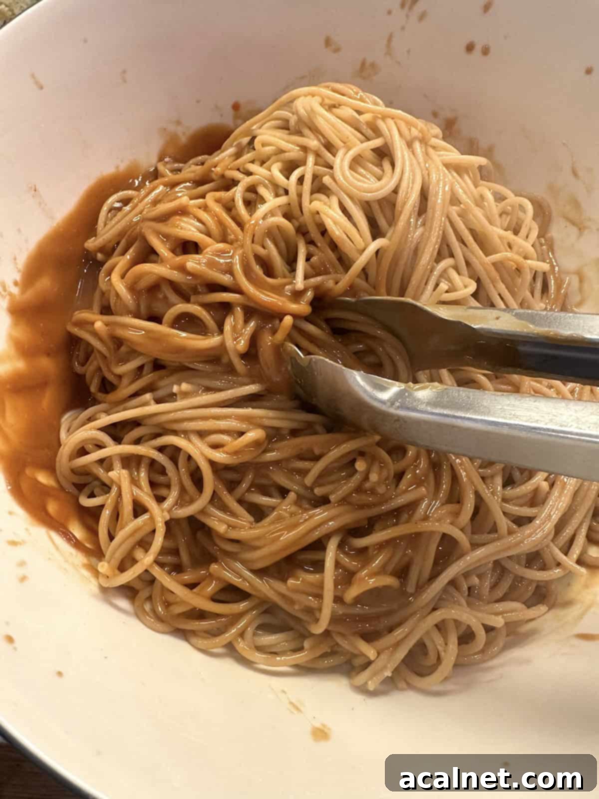 Cooked spaghetti noodles in a bowl with sauce and tongs.