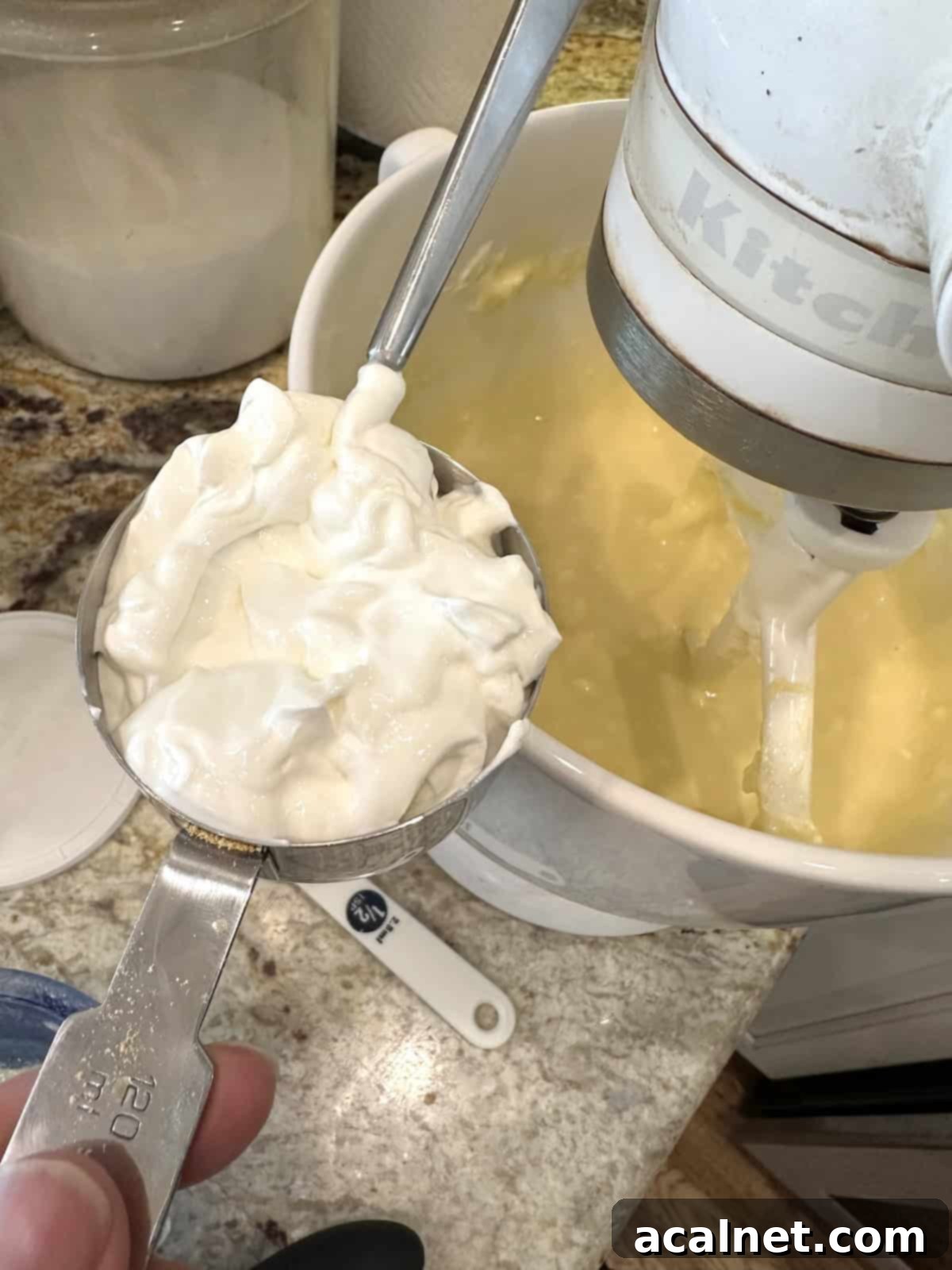 A measuring scoop filled with creamy sour cream rests near a large mixing bowl containing cream cheese, sugar, and other cheesecake filling ingredients, ready to be blended.