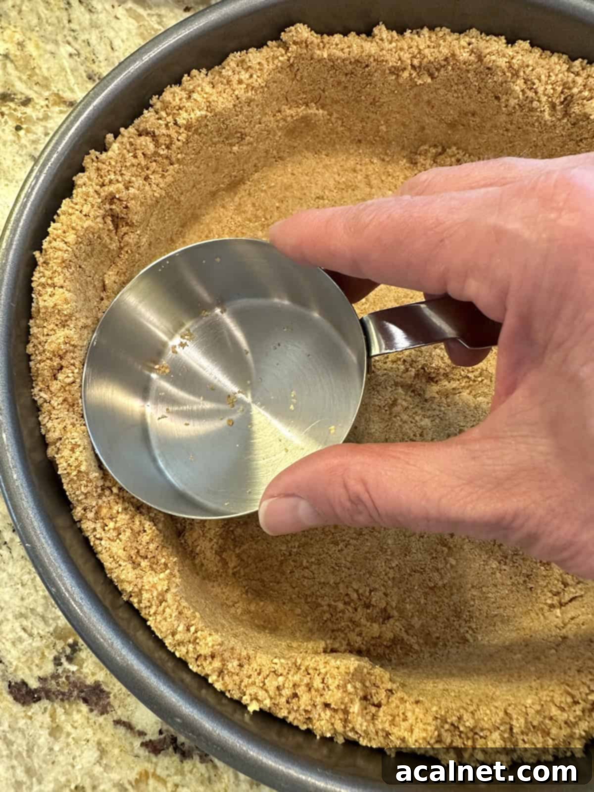 An empty glass measuring cup is used to firmly press and flatten the graham cracker crust mixture evenly across the bottom and up the sides of a 9-inch springform pan.