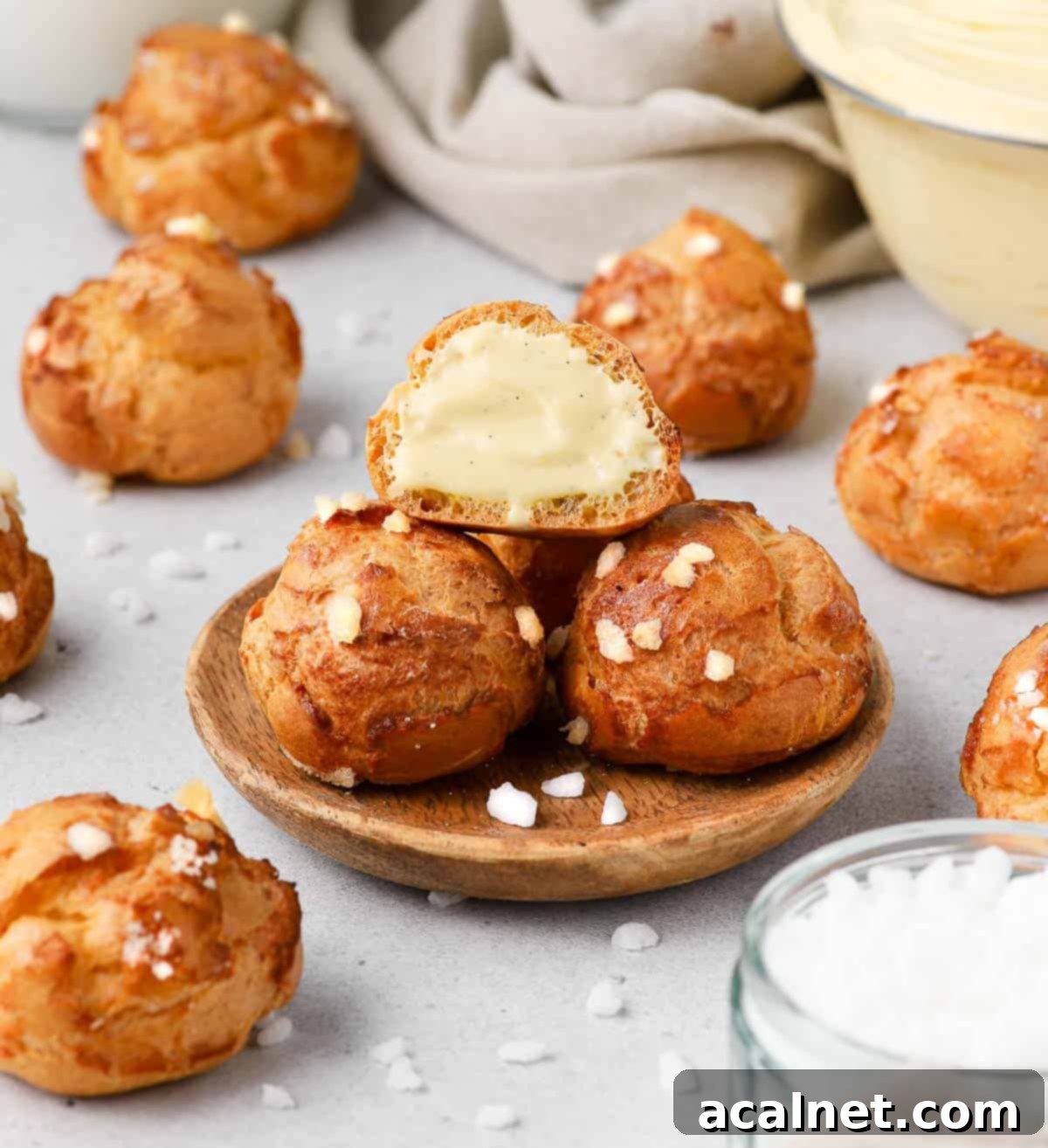 Three golden-brown chouquettes stacked on a small wooden plate, with one cut in half revealing a creamy filling.