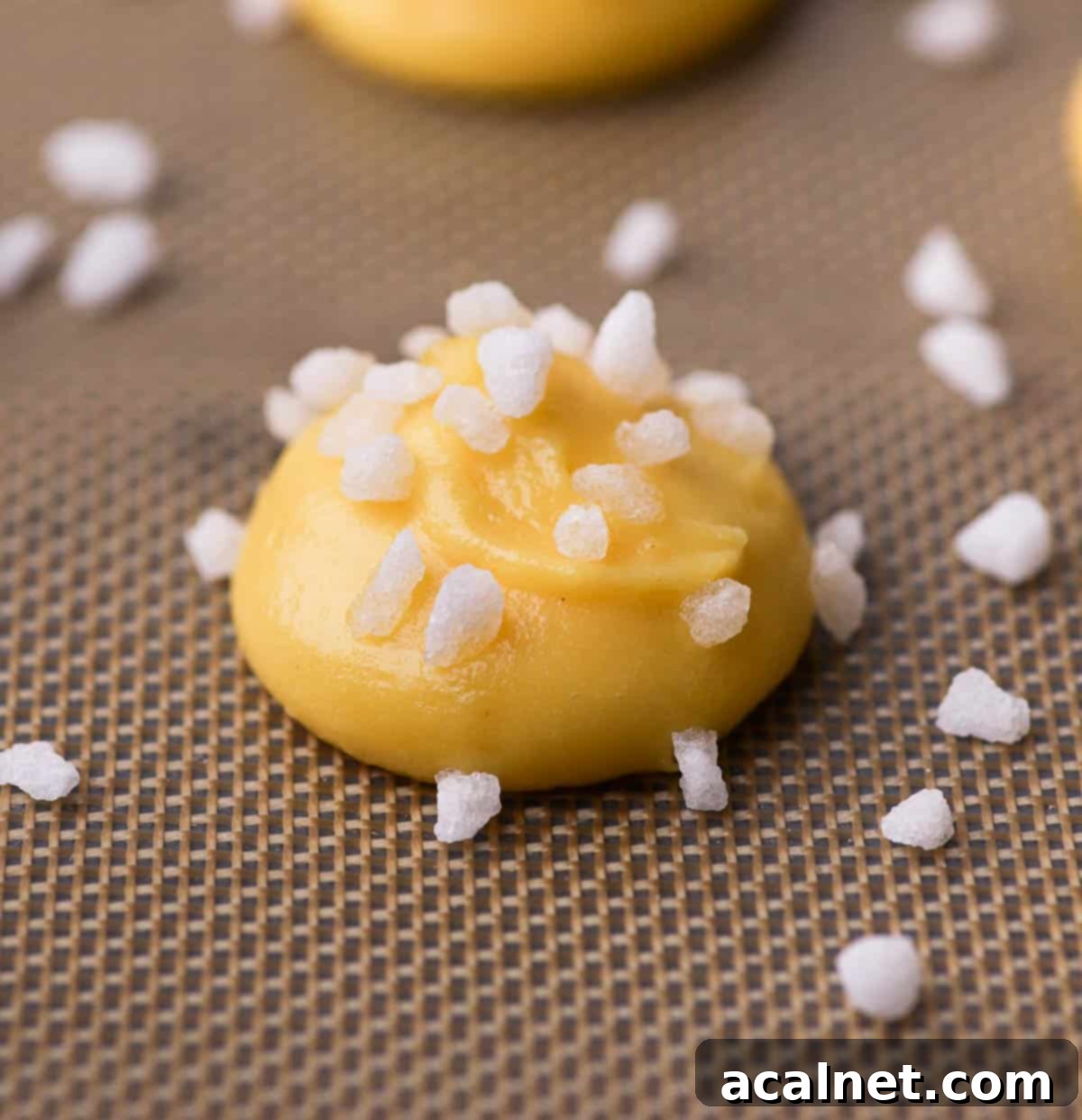 An unbaked chouquette, perfectly piped onto a baking mat and generously topped with pearl sugar, ready for the oven.