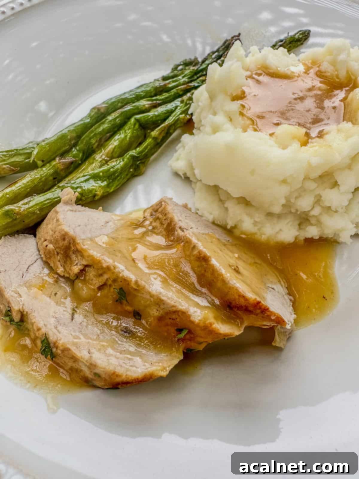 A serving of sliced pork tenderloin with orange sauce on a plate with mashed potatoes and asparagus.