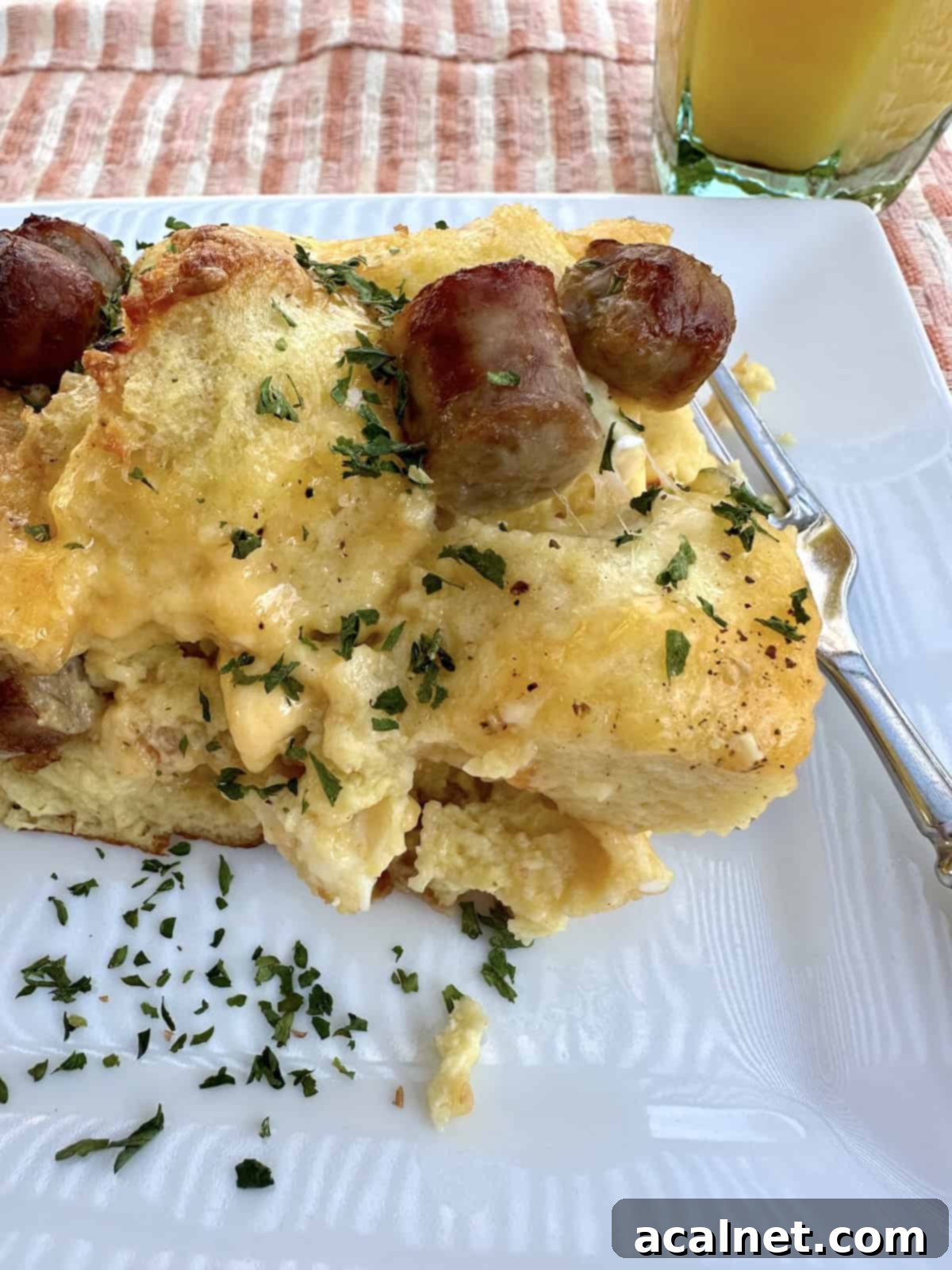 Cheesy Sausage and Egg Bake 2 A generous serving of golden-brown cheese, egg, and sausage casserole on a white plate, accompanied by a fork and a refreshing glass of orange juice. The casserole shows layers of bread, melted cheese, and crumbled sausage.