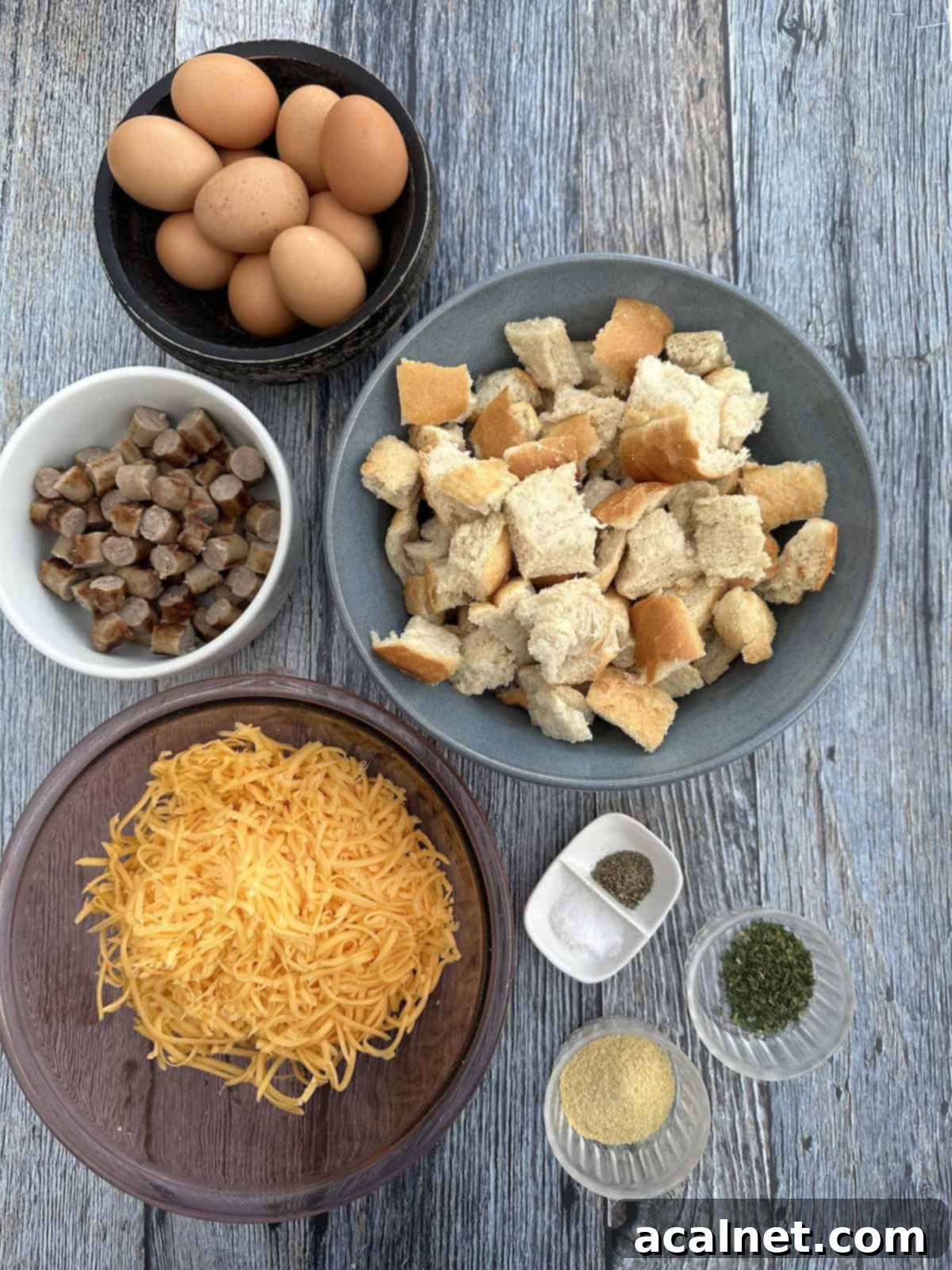 Cheesy Sausage and Egg Bake 4 Various ingredients for the sausage casserole neatly arranged in bowls on a rustic wooden table, including shredded cheese, raw eggs, milk, cooked sausage, and cubed bread.