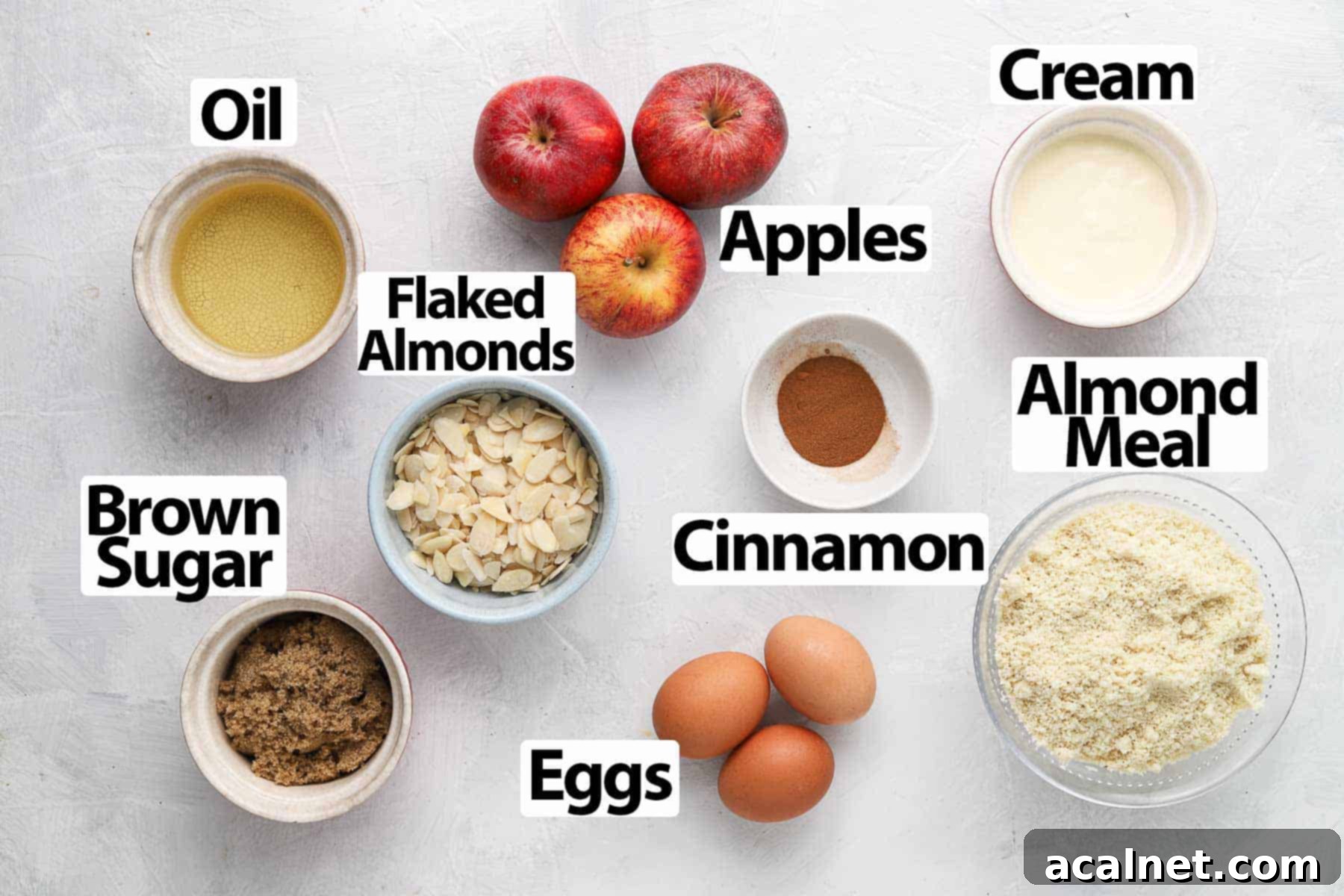 Heavenly Gluten-Free Apple Almond Cake 3 A collection of fresh ingredients for apple almond cake, including apples, eggs, almond meal, brown sugar, cream, oil, and cinnamon, neatly arranged on a white surface.
