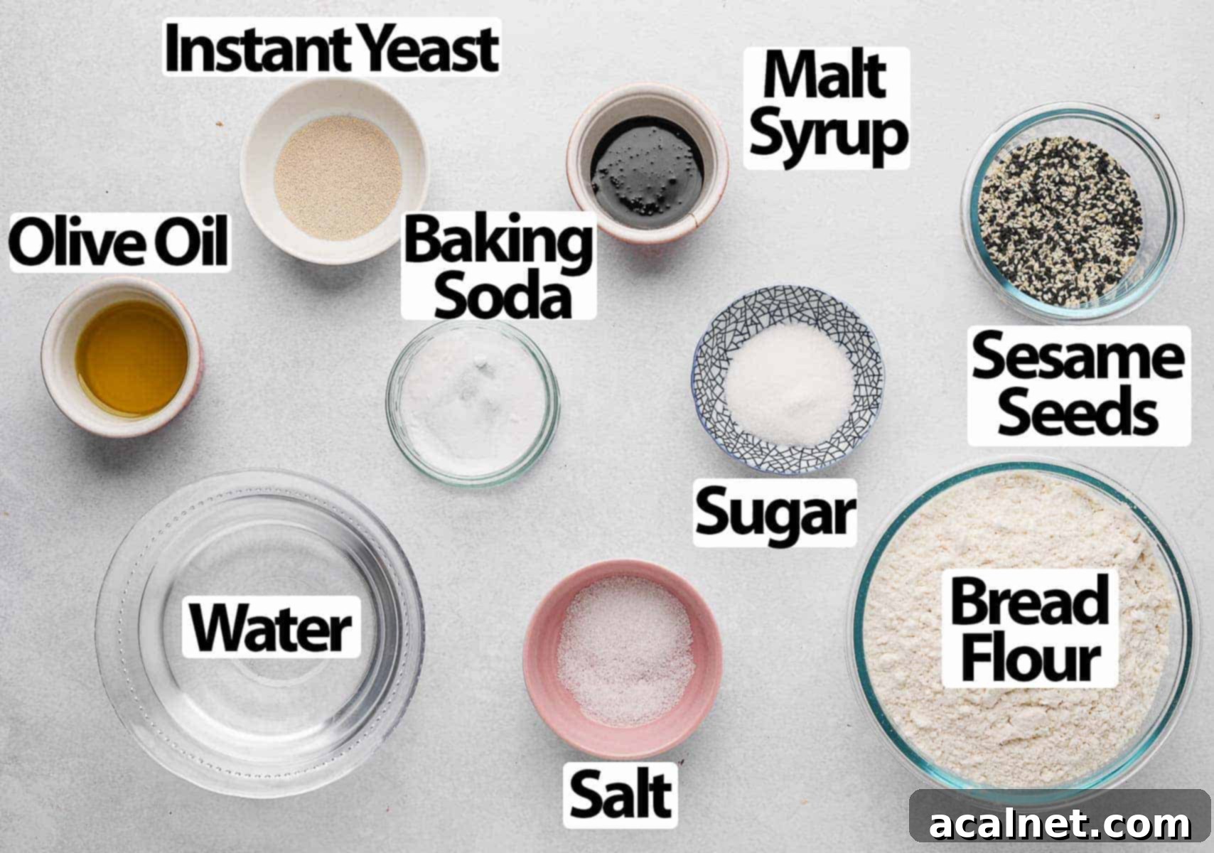 A selection of bagel ingredients laid out on a grey surface, including flour, yeast, oil, and sesame seeds.