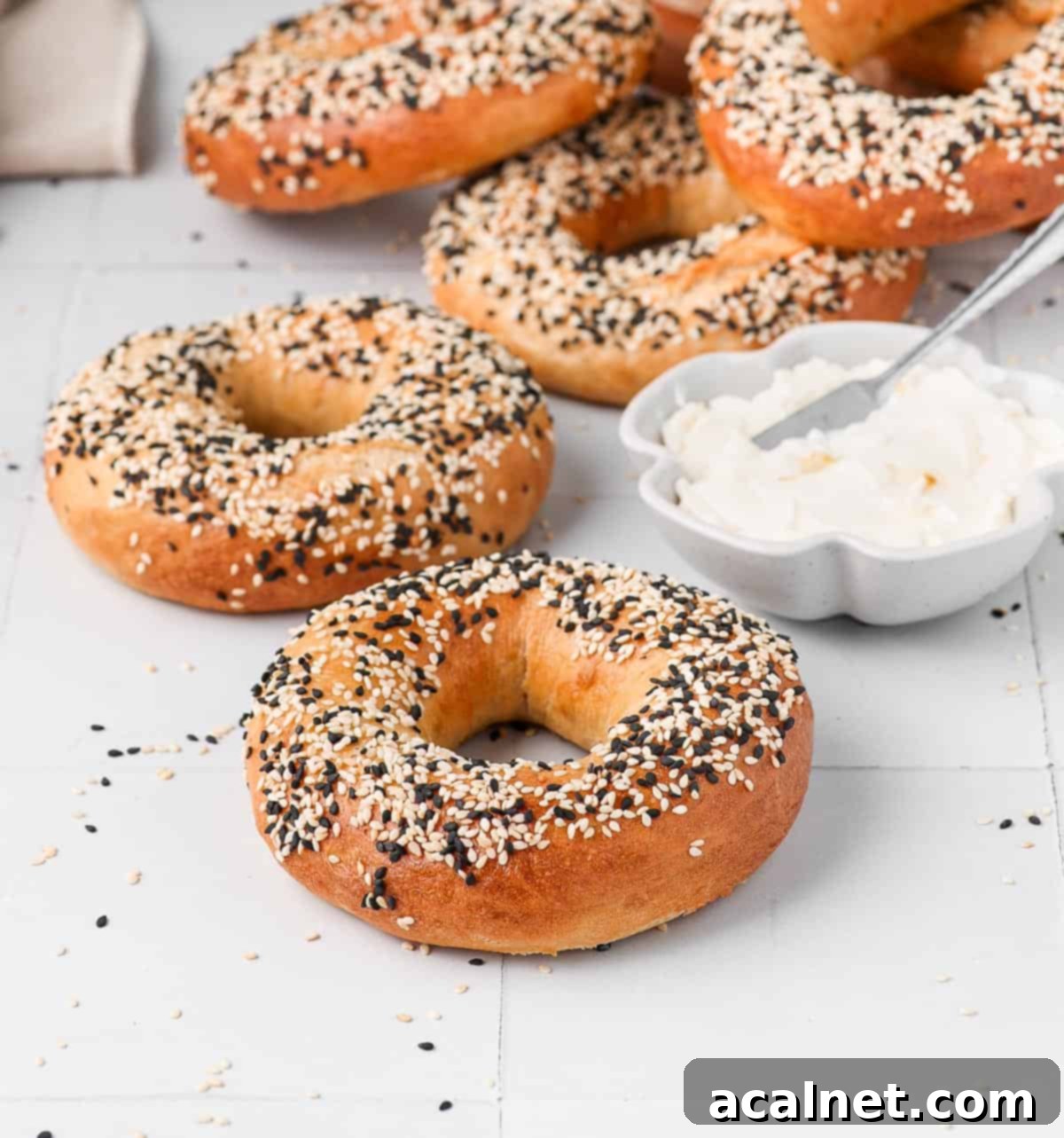 Freshly baked homemade sesame bagels on a white surface, with a bowl of cream cheese nearby.