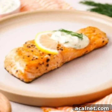A portion of perfectly cooked salmon on a plate, generously topped with a creamy dill sauce and garnished with a lemon slice.