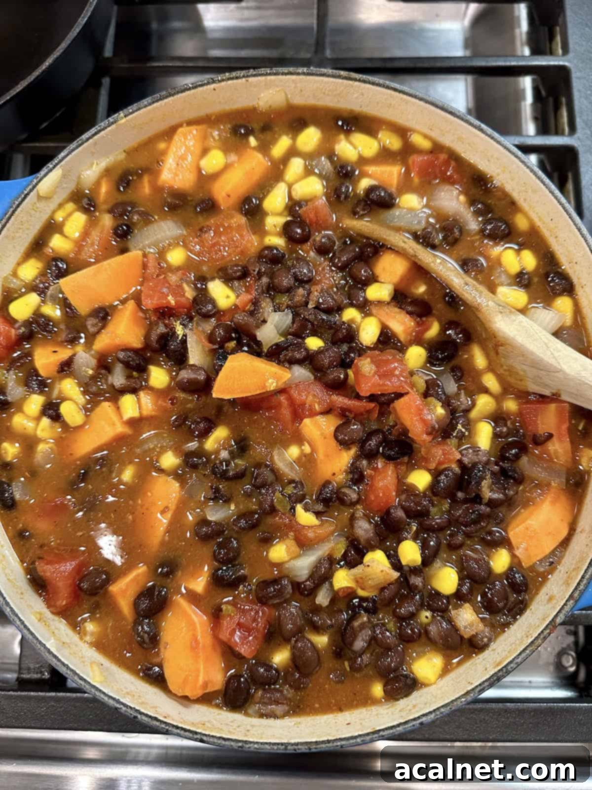A large pot of vibrant black bean soup with sweet potatoes and corn, ready to be served, on a rustic wooden surface with a ladle.