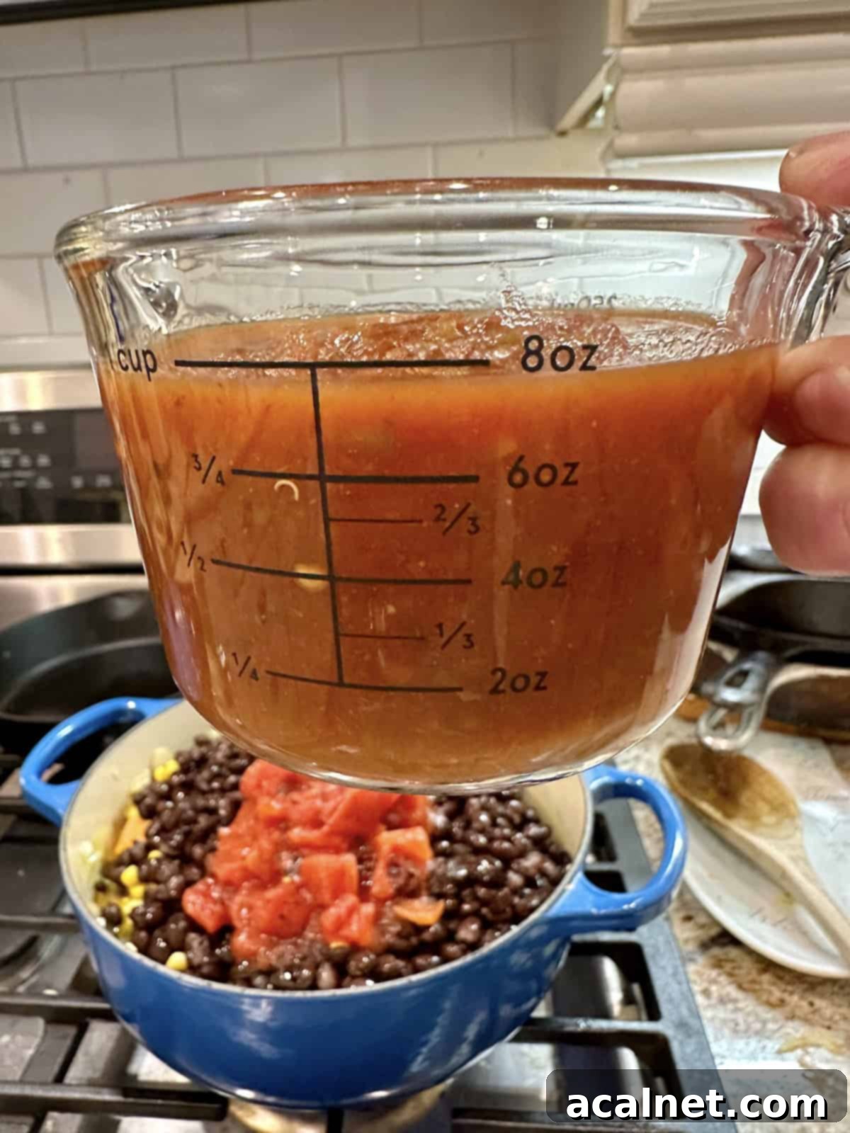 A cup of vibrant medium salsa being poured into a large pot containing black beans, corn, sweet potatoes, and onions, adding a burst of flavor.
