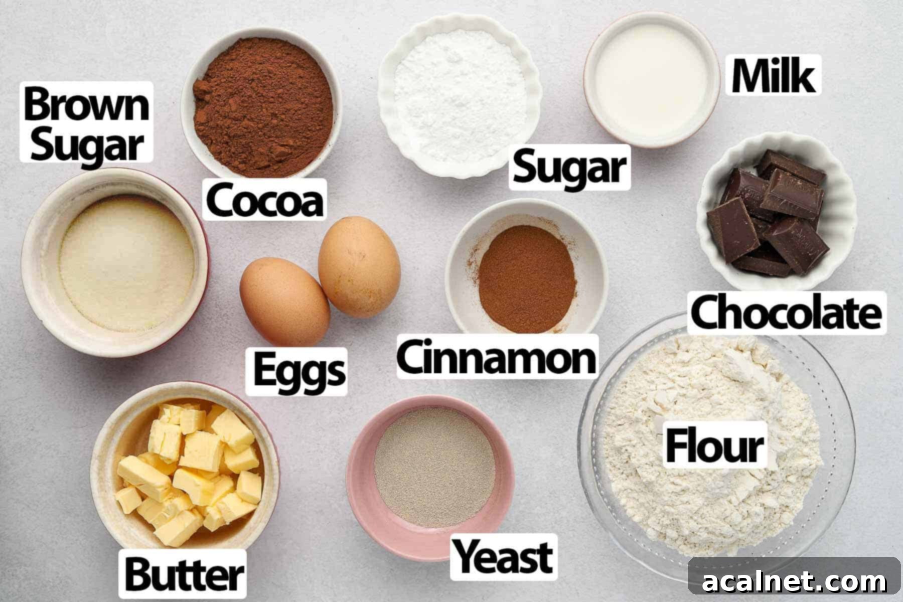 A flat lay of ingredients for triple chocolate cinnamon rolls, including milk, flour, cocoa powder, butter, eggs, sugar, yeast, cinnamon, and dark chocolate chunks.