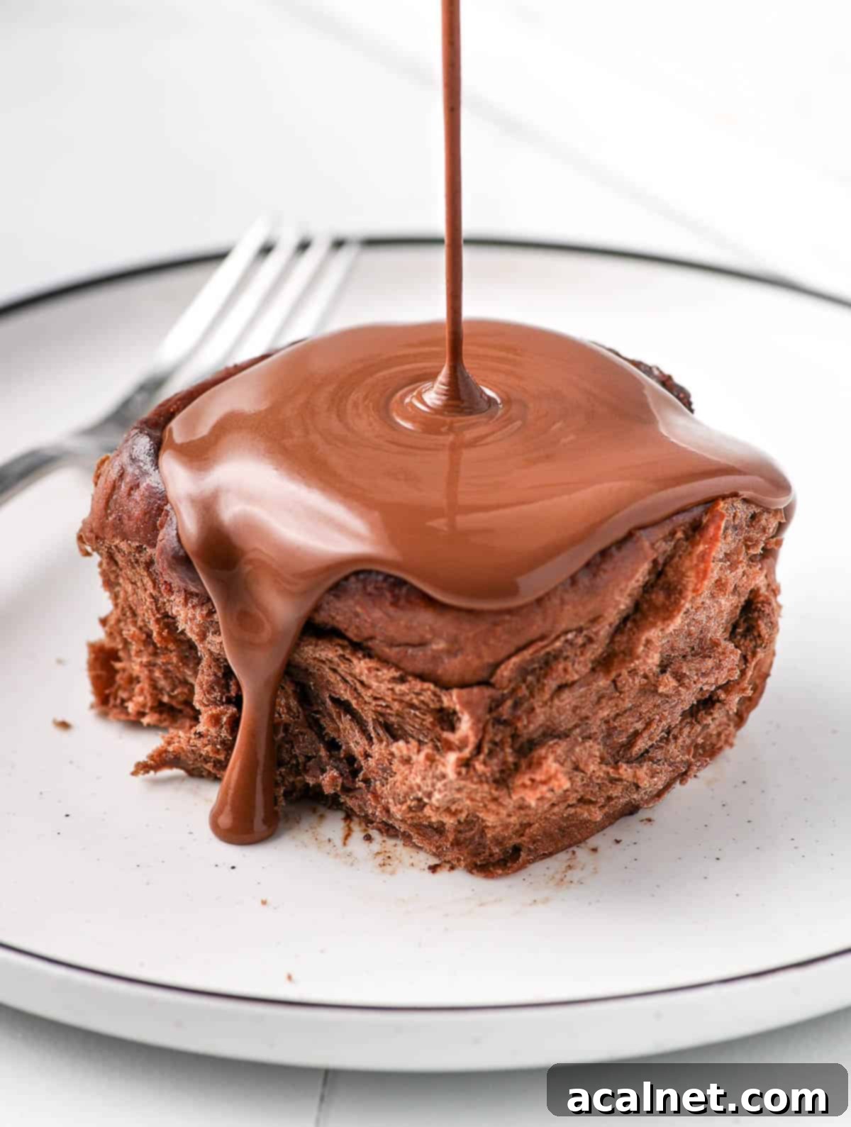 A close-up shot of warm dark chocolate ganache being poured generously over a single triple chocolate cinnamon roll on a white plate.