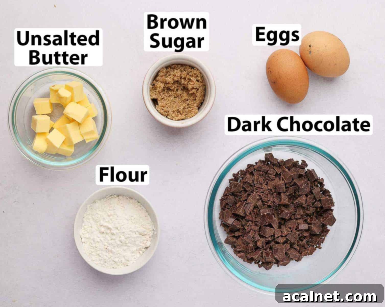 The five core ingredients for chocolate fondant cakes laid out on a grey surface: dark chocolate, butter, eggs, brown sugar, and flour.