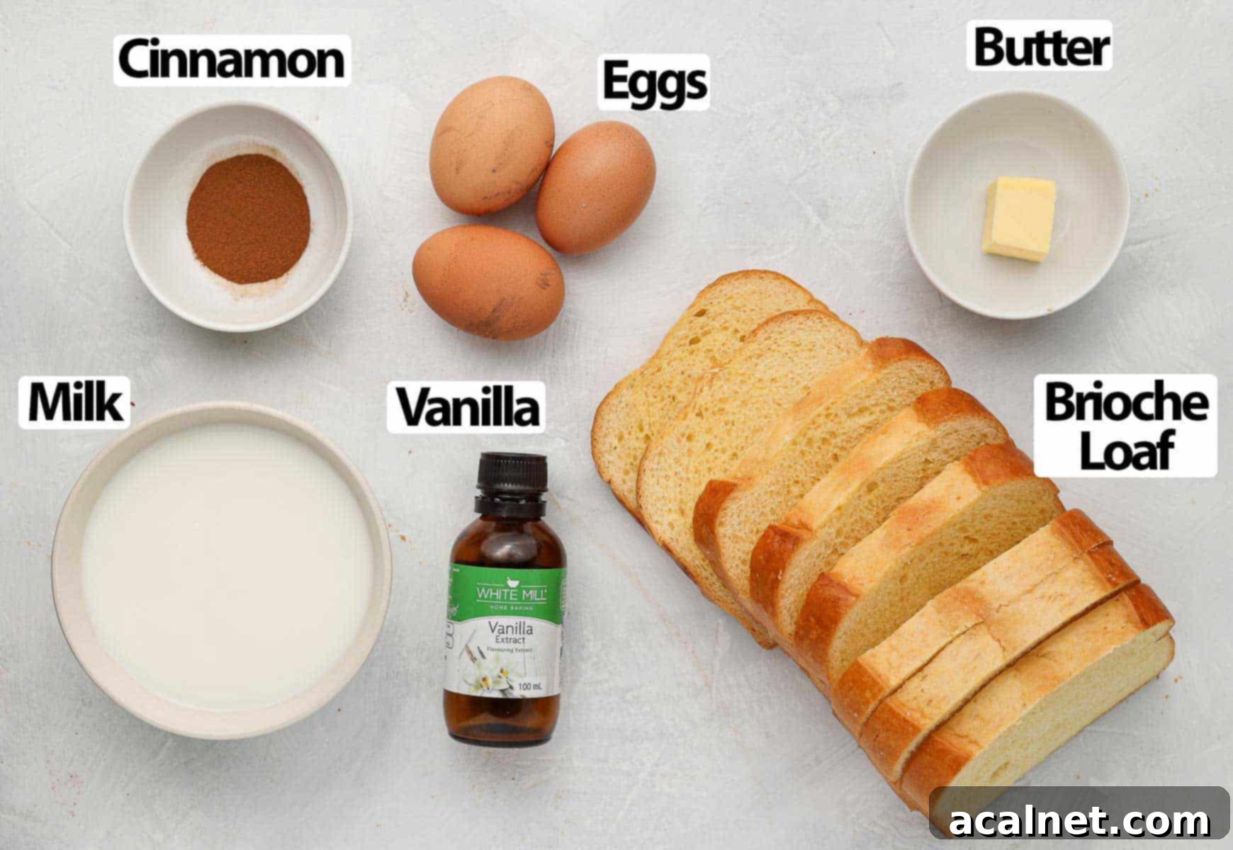 Collection of fresh ingredients for brioche French toast, including eggs, milk, cinnamon, vanilla, butter, and brioche bread slices, artfully arranged on a white surface.
