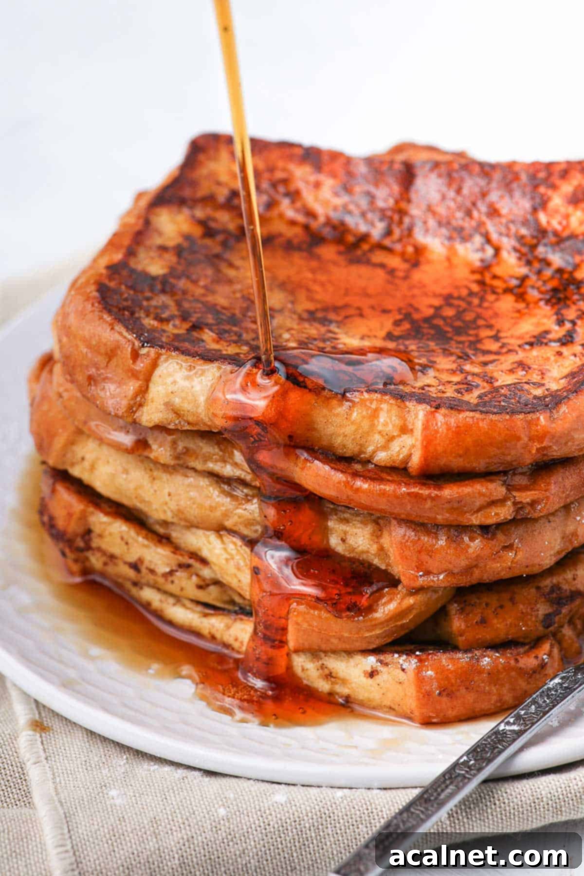 Rich maple syrup being drizzled over a beautifully stacked tower of brioche French toast, highlighting its golden hue and fluffy texture.