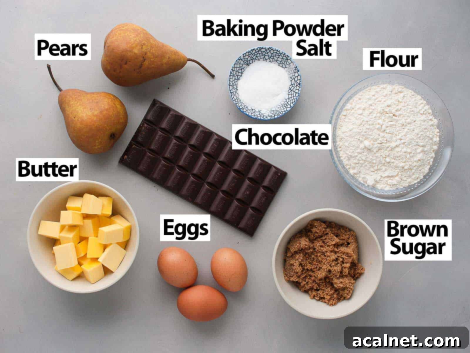 A collection of fresh ingredients laid out on a grey surface, including dark chocolate, butter, eggs, flour, pears, and sugar, ready for baking this delightful cake.