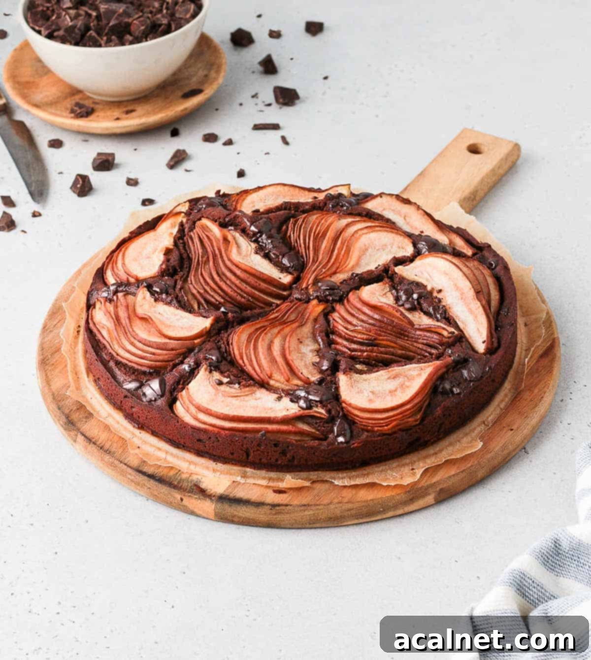 A whole Chocolate Pear Cake on a round wooden board, garnished with additional chocolate chunks, ready for serving.