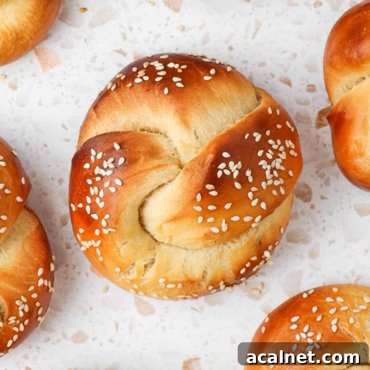 A single perfectly golden mini challah roll, topped with sesame seeds, resting on a white and pink terrazzo surface, ready to be enjoyed.