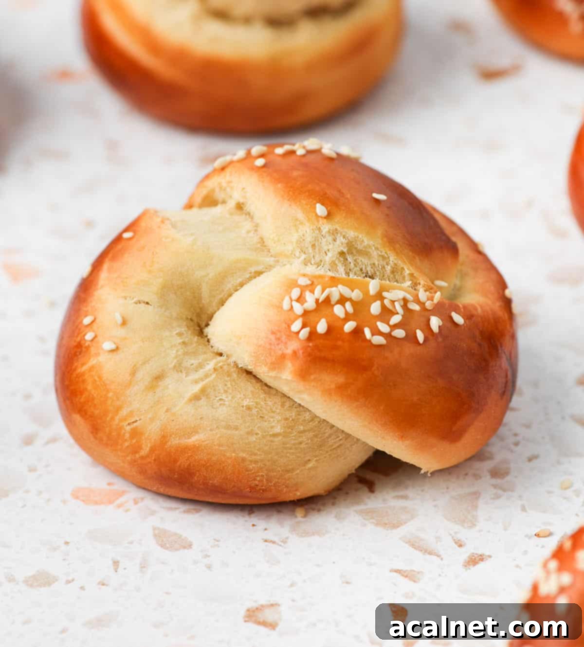 Close-up shot of a single perfectly baked mini challah roll with sesame seeds on top, resting on a white and pink terrazzo surface, highlighting its golden crust and soft texture.