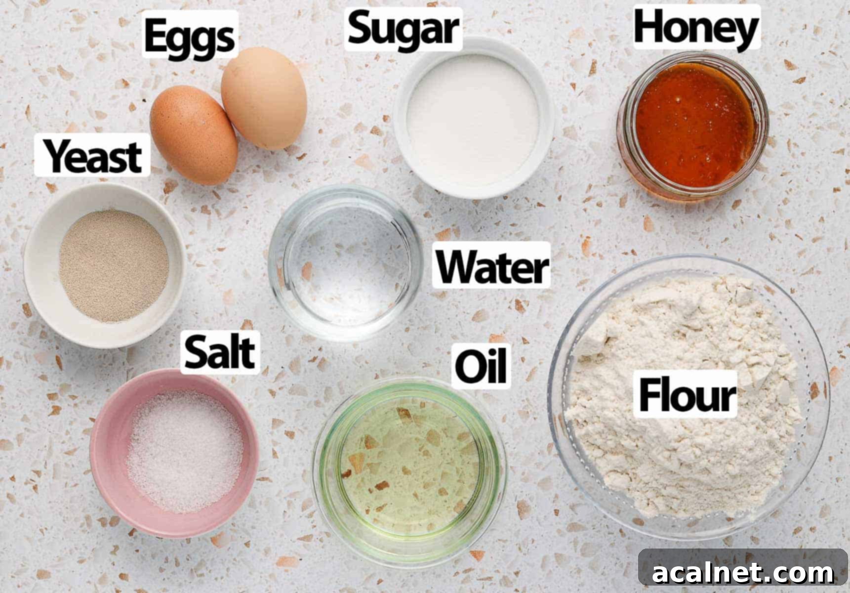 A collection of fresh ingredients for baking challah rolls, including flour, eggs, yeast, honey, and oil, neatly arranged on a white and pink terrazzo surface.