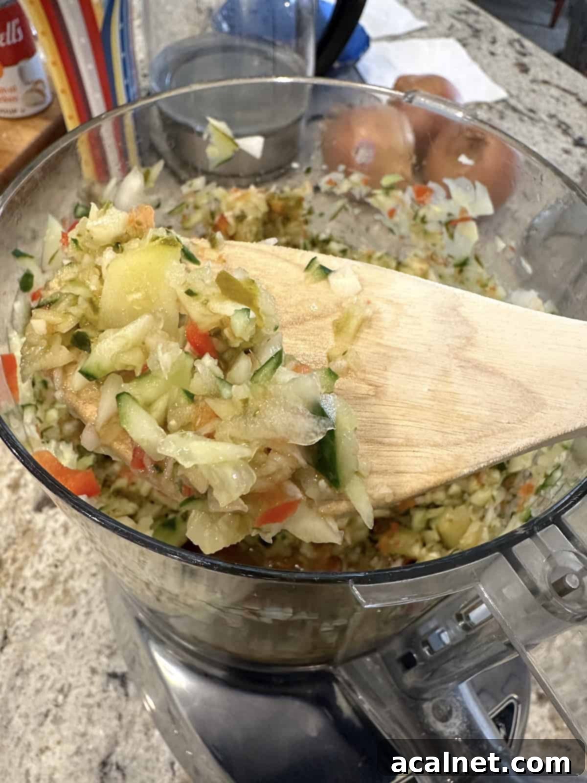 A wooden spoon scooping out some of the chopped pickles and cucumbers from a food processor bowl.