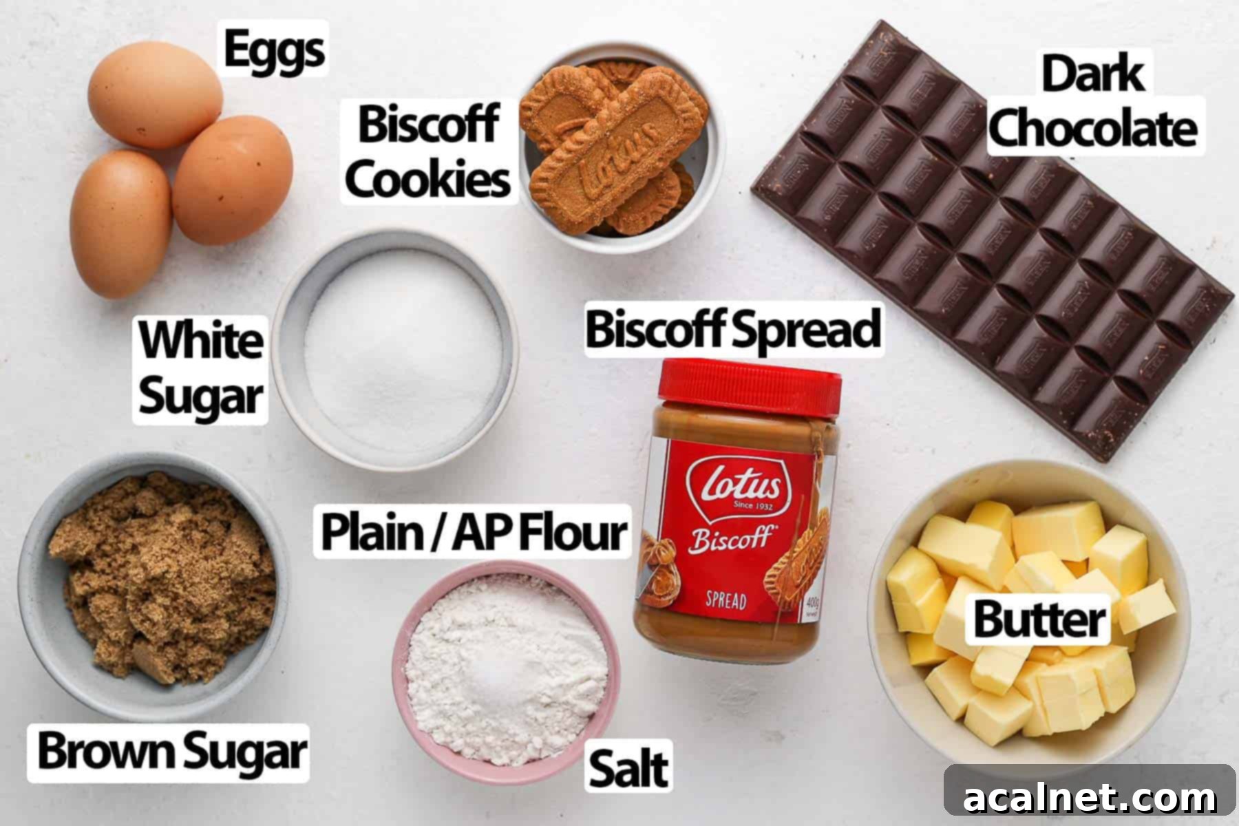 A carefully arranged collection of ingredients for Biscoff brownies, including chopped dark chocolate, sticks of butter, jars of Biscoff spread, fresh eggs, and bowls of brown and white sugar, all set on a pristine white surface.