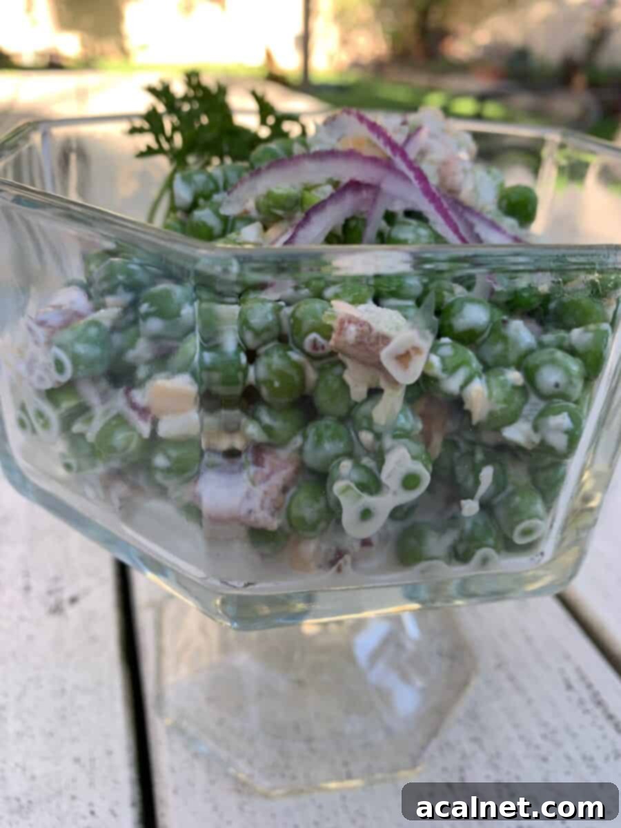 Pea salad in a clear glass serving bowl, garnished with a sprig of parsley, on an outdoor wooden table with a vintage-style white pitcher.