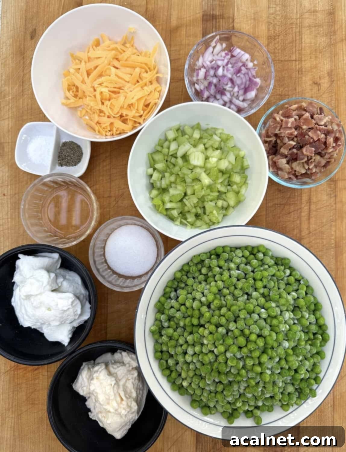 Bowls of ingredients for pea salad including frozen peas, chopped celery, diced red onion, shredded cheddar cheese, and a bowl of crispy bacon bits, arranged on a wooden cutting board.