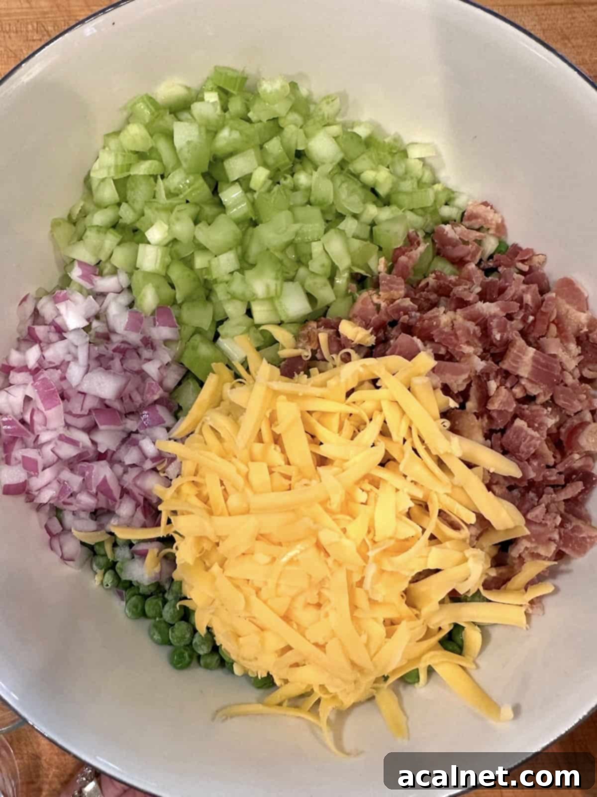 A large glass bowl filled with thawed frozen peas, topped with piles of shredded cheddar cheese, diced red onion, crispy bacon bits, and chopped celery, ready to be mixed.